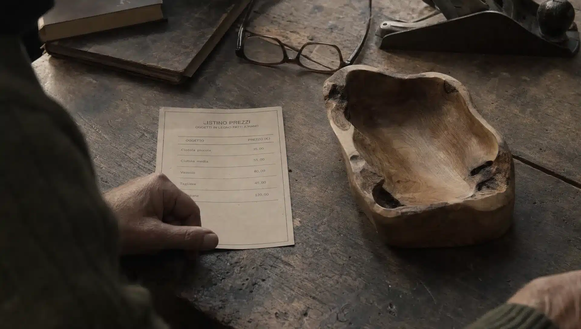 Su un tavolo di lavoro, un listino prezzi resta accanto a un oggetto artigianale finito mentre la scelta si sposta dal numero al valore.