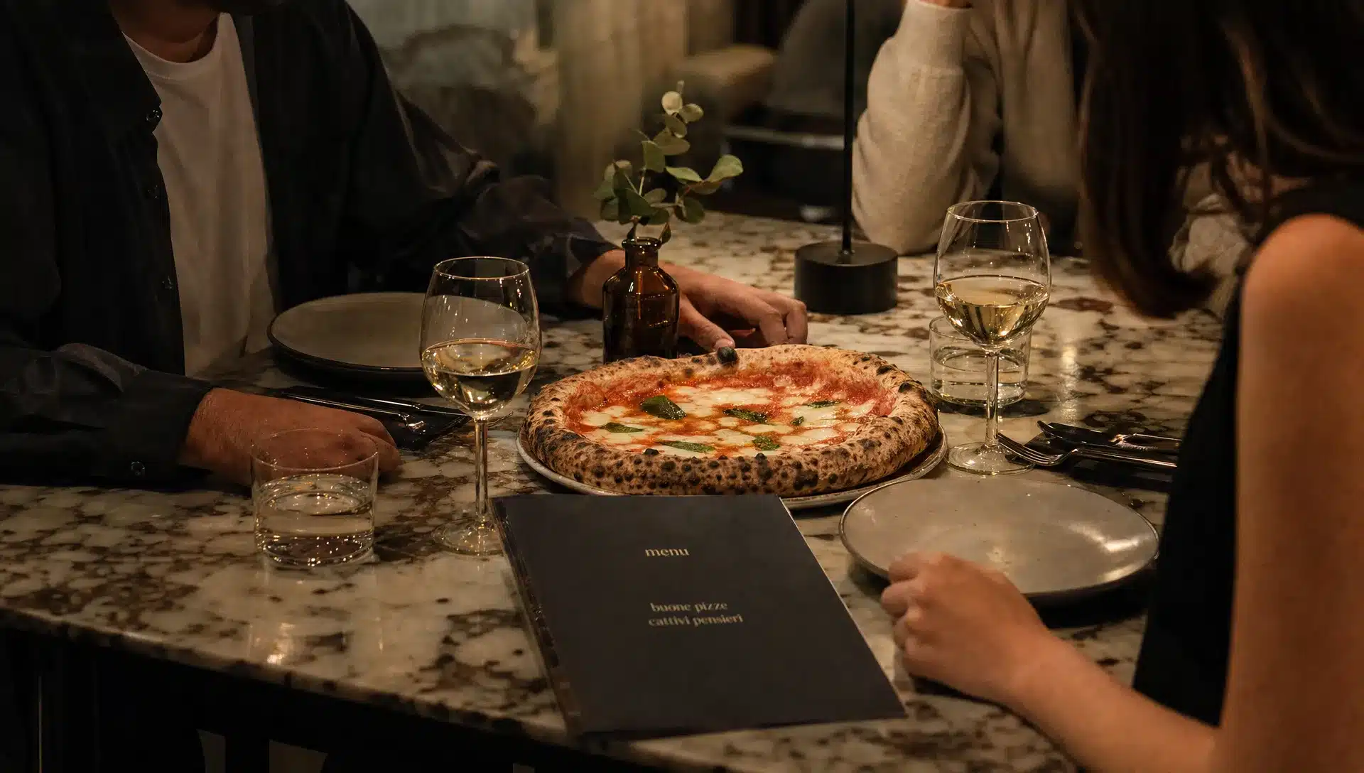 Un tavolo apparecchiato per una serata condivisa in pizzeria, con una pizza appena servita al centro e il menu chiuso vicino ai piatti e ai bicchieri.